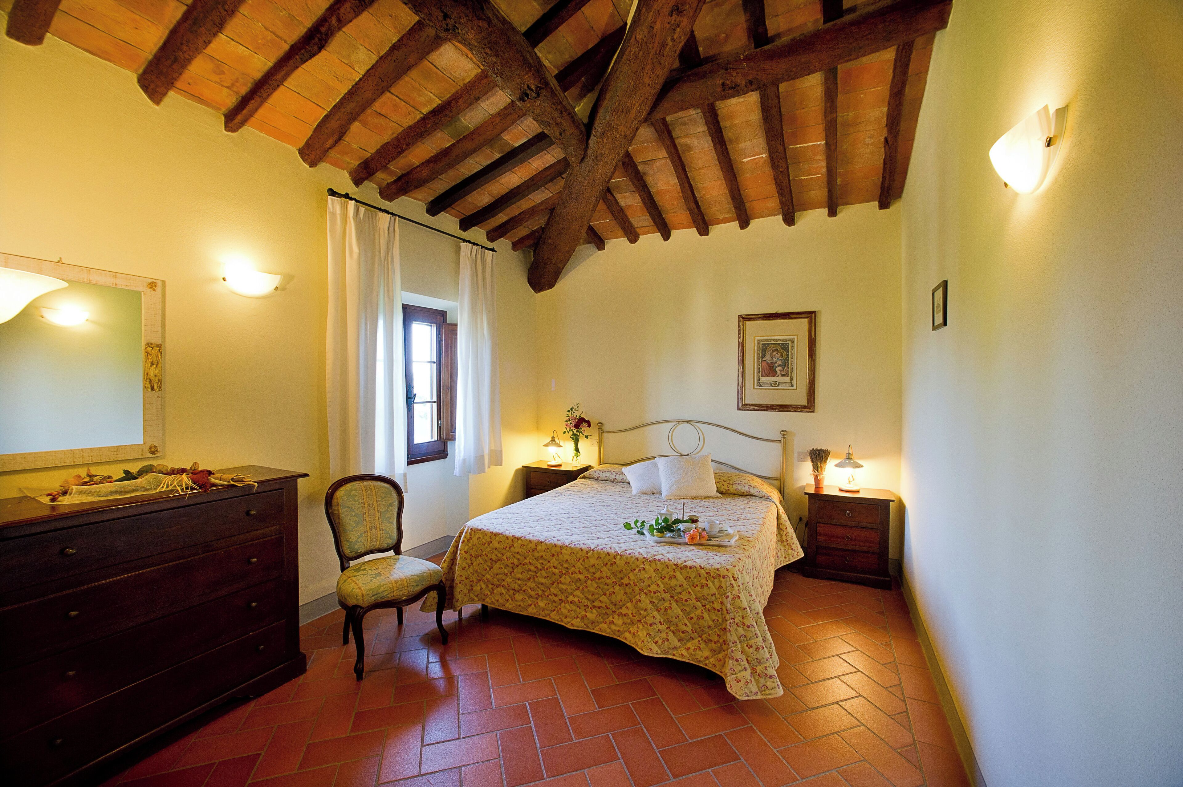 A bedroom with a wooden ceiling, a bed with a floral bedspread, a dresser, a chair, and a nightstand with a lamp.