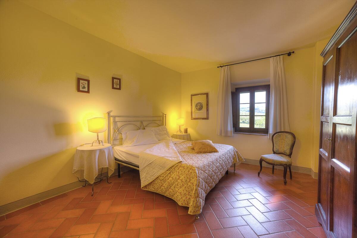 A bedroom with a bed, two bedside tables, a chair, a window with curtains, and a door.