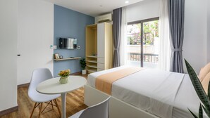 A modern hotel room with a bed, a desk, a chair, a small table with a vase of flowers, a wardrobe, and a window with curtains. - Cozrum Homes - Summer's House (Ho Chi Minh City)