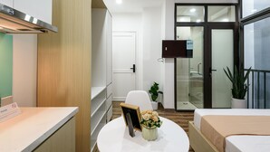 A modern hotel room with a bed, a small table with a vase, a chair, a TV mounted on the wall, and a sliding glass door leading to a bathroom. - Cozrum Homes - Summer's House (Ho Chi Minh City)