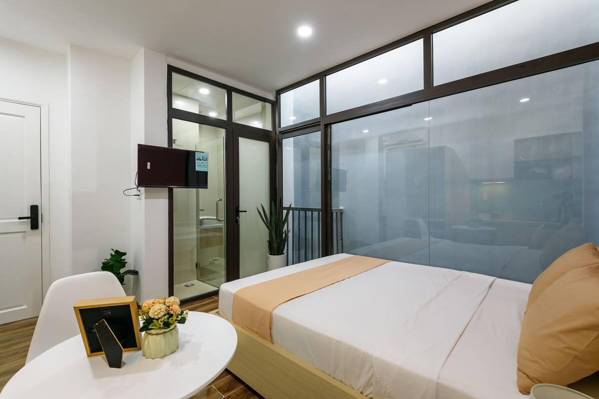 Una habitación de hotel moderna con una cama grande, una mesita pequeña con un marco de fotos y flores, una puerta blanca y un ventanal con vistas al exterior.
