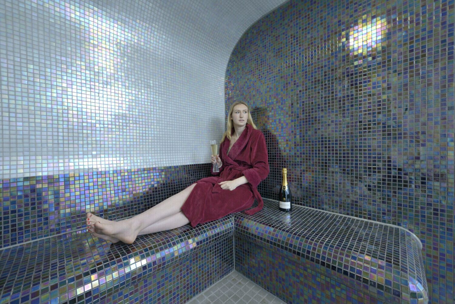 A woman in a red robe sitting on a bench in a room with mosaic tile walls and a bottle of champagne.