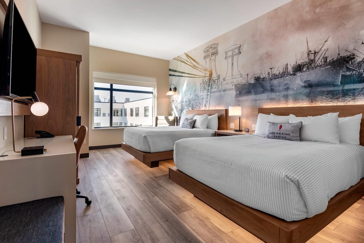 Une chambre d’hôtel avec deux lits, un bureau et une grande fresque représentant un paysage portuaire.