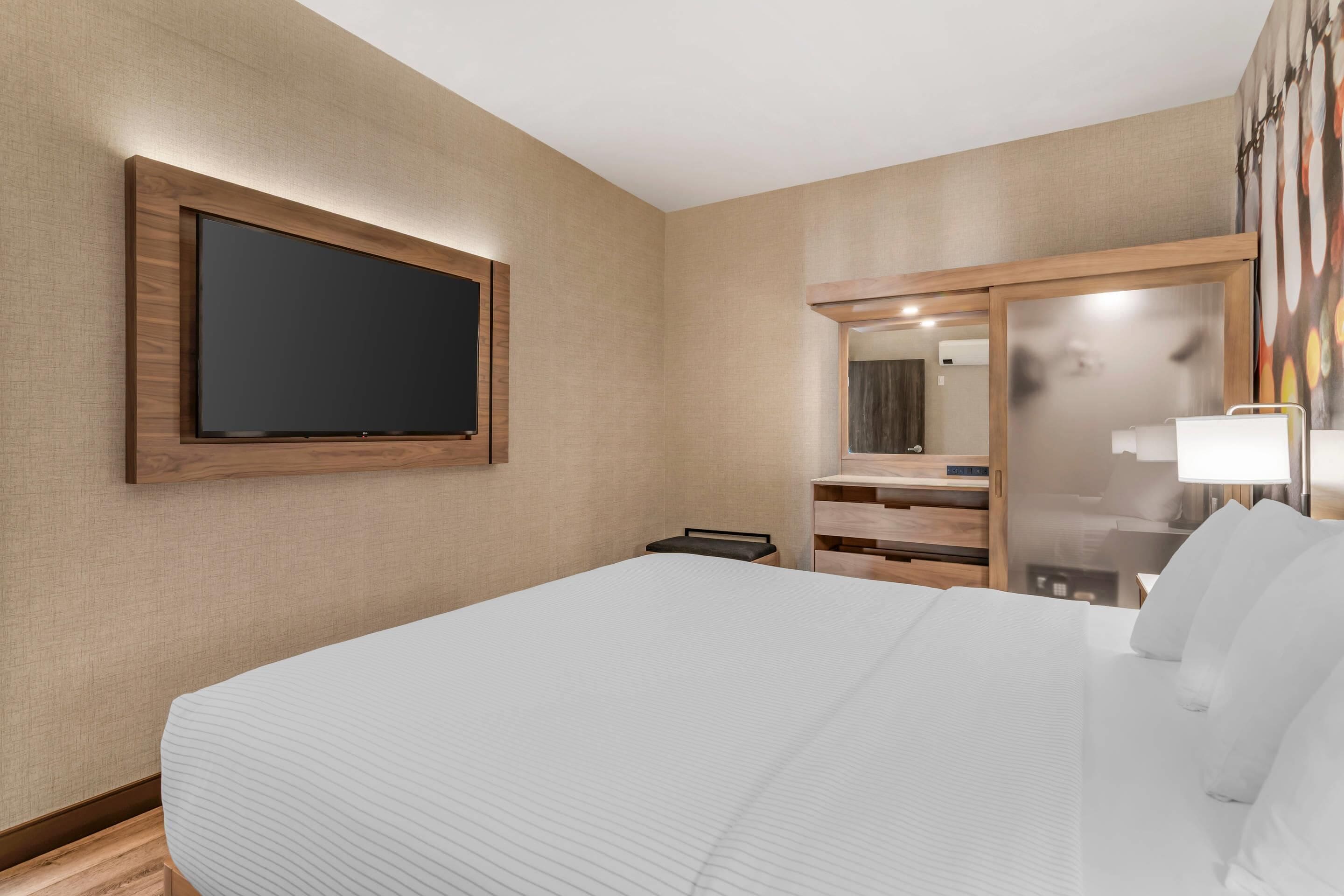 Una habitación de hotel con una cama grande, un televisor de pantalla plana montado en la pared, un espejo y una mesita de noche con una lámpara.