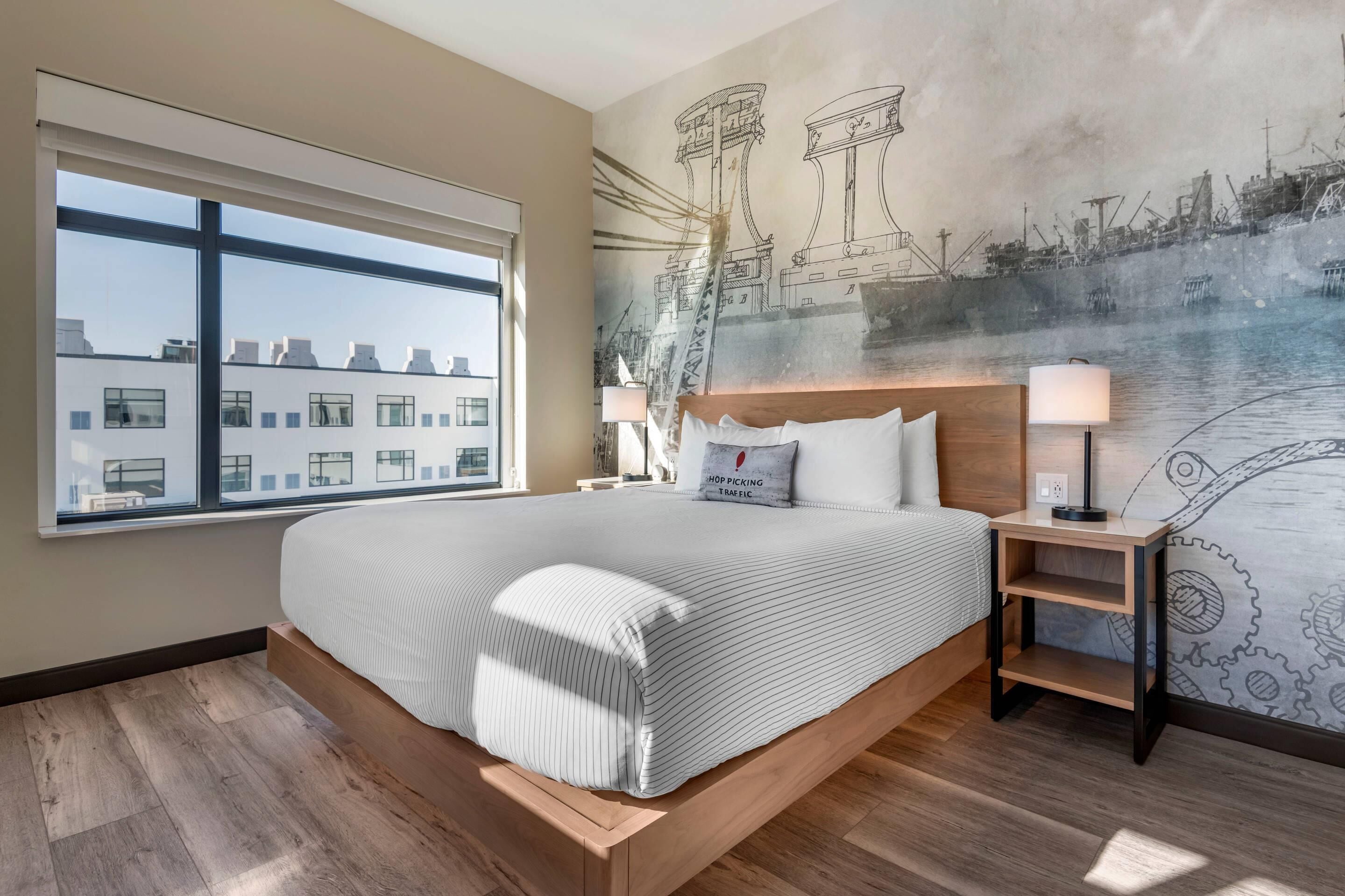 Un hotel moderno con una cama grande, vistas de la ciudad y un mural con grúas y barcos en la pared.