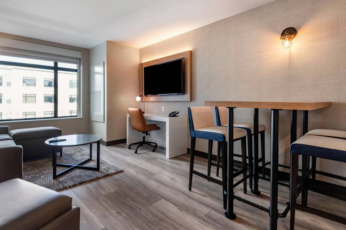 Une chambre d’hôtel moderne dotée d’une grande fenêtre, d’un téléviseur à écran plat fixé au mur, d’un bureau avec une chaise et d’un coin salon avec des tabourets.