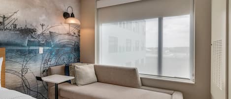 A modern hotel room with a beige sofa, a small table, a wall mural of ships, and a large window with a view of buildings.