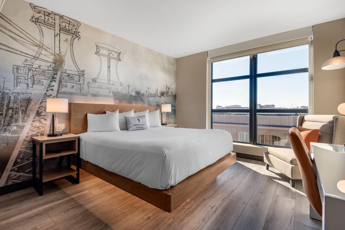 Une chambre d’hôtel moderne, dotée d’un grand lit, d’un bureau et d’une chaise. La chambre dispose d’une grande fenêtre offrant une vue sur la ville.