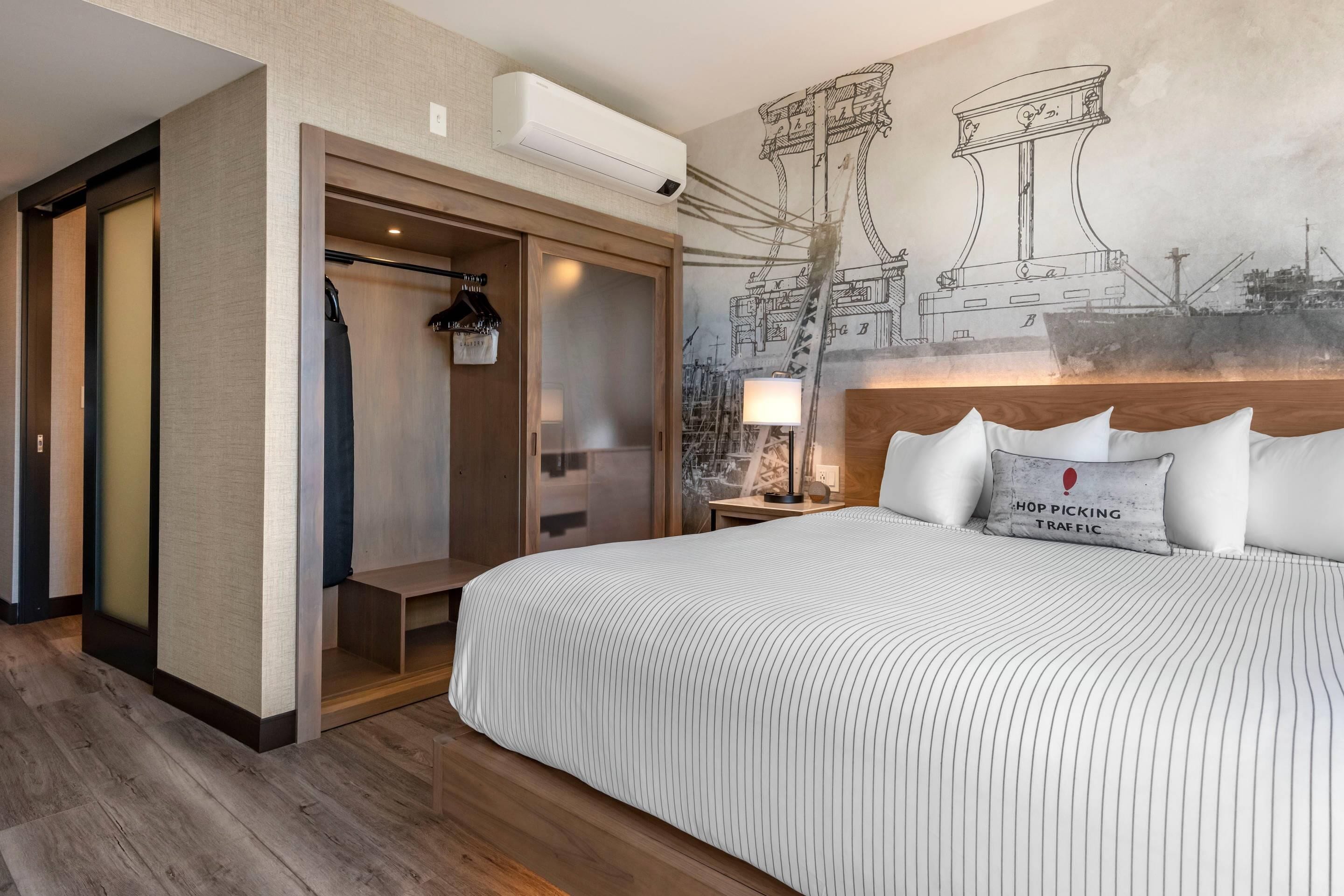 Habitación de hotel con una cama grande, un vestidor, un mural en la pared y una mesita de noche con lámpara.