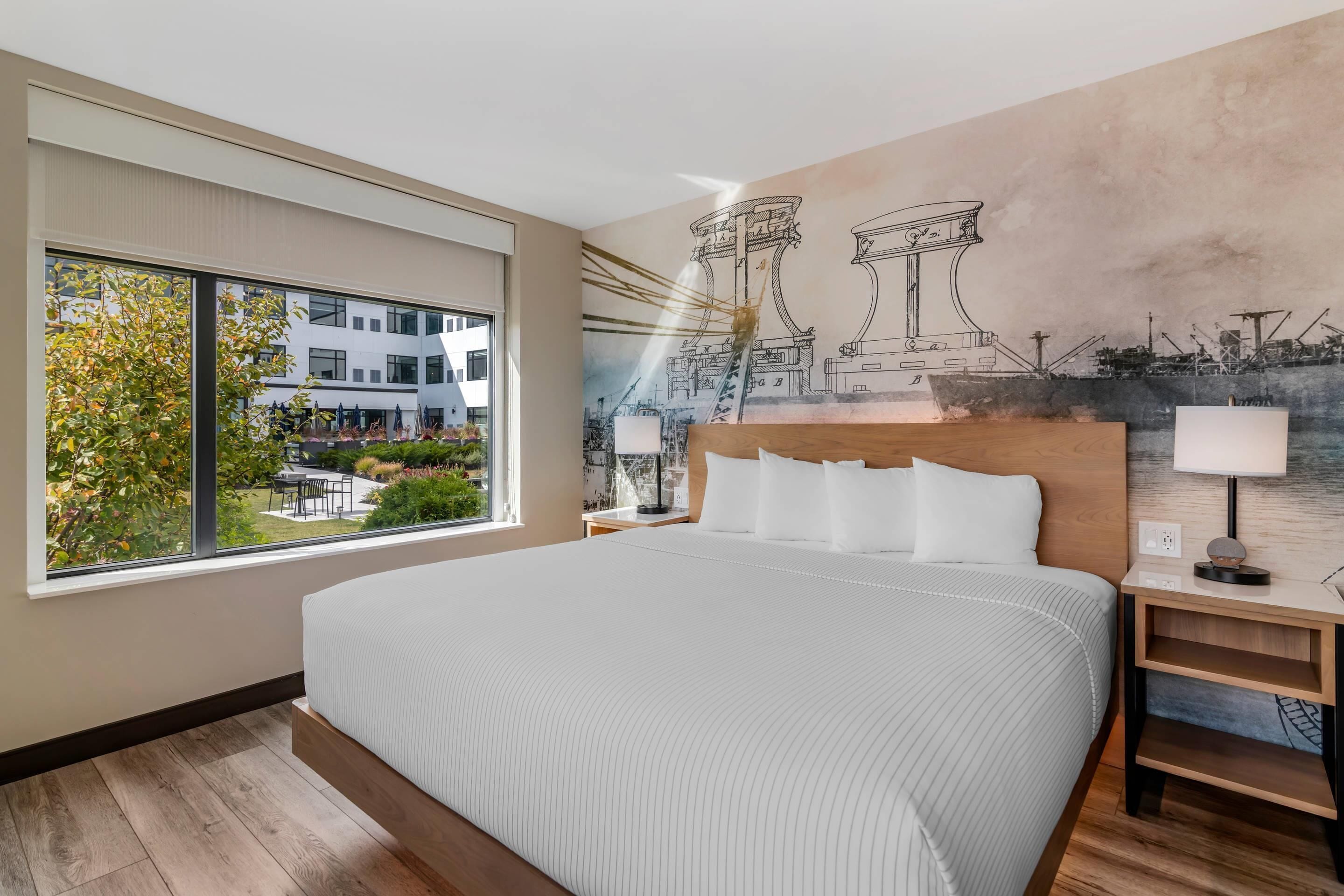A modern hotel room with a large bed, a view of the outside, and a mural of architectural structures.