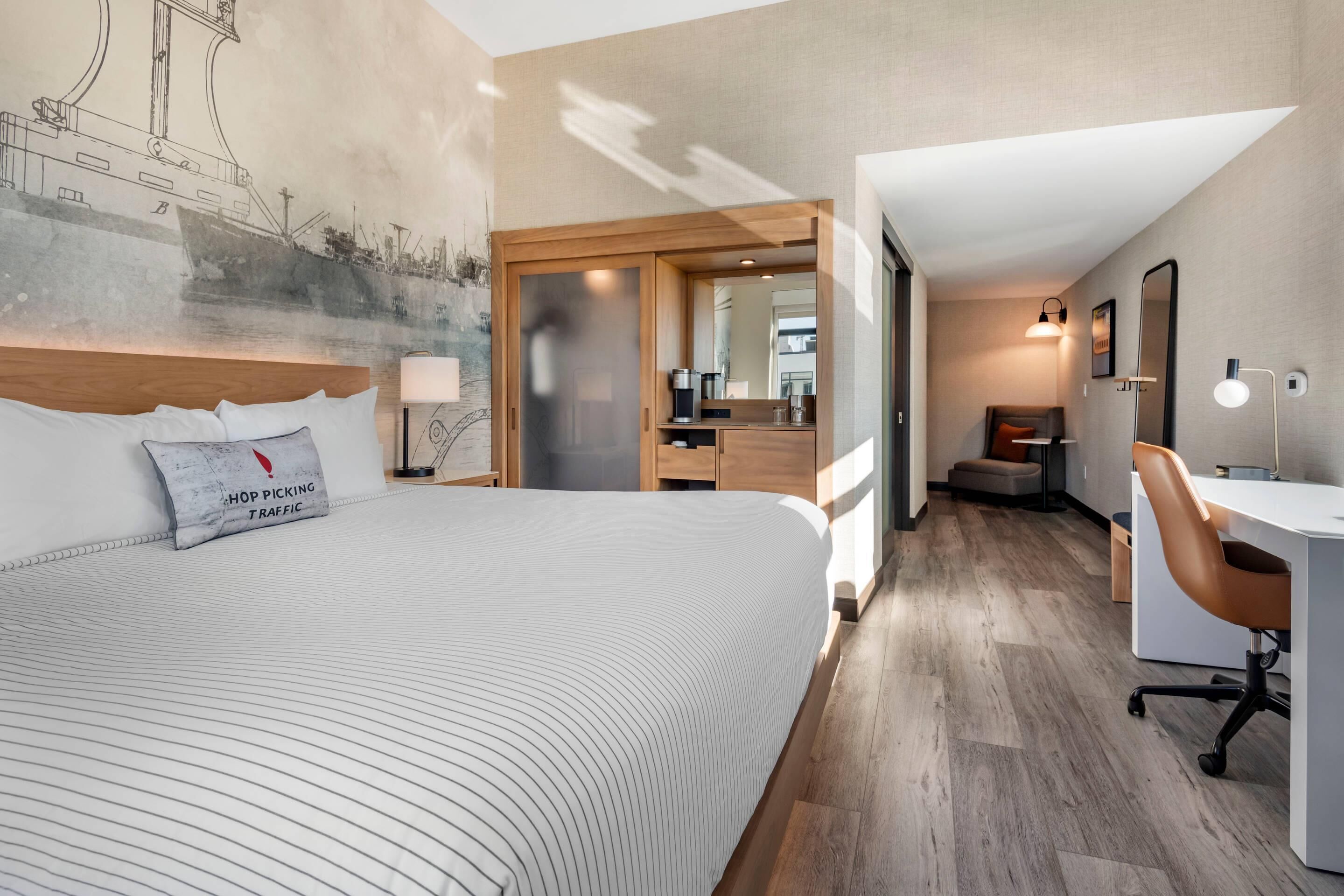 A modern hotel room with a large bed, a desk, and a chair. There is a large window and a wall mural.