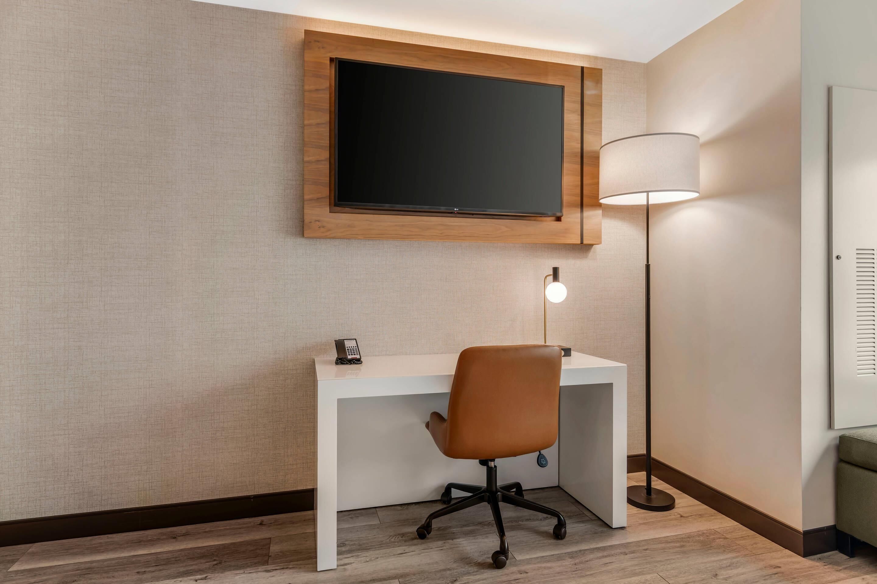 A hotel room with a white desk, a brown office chair, a mounted flat-screen TV, a floor lamp, and a small telephone.
