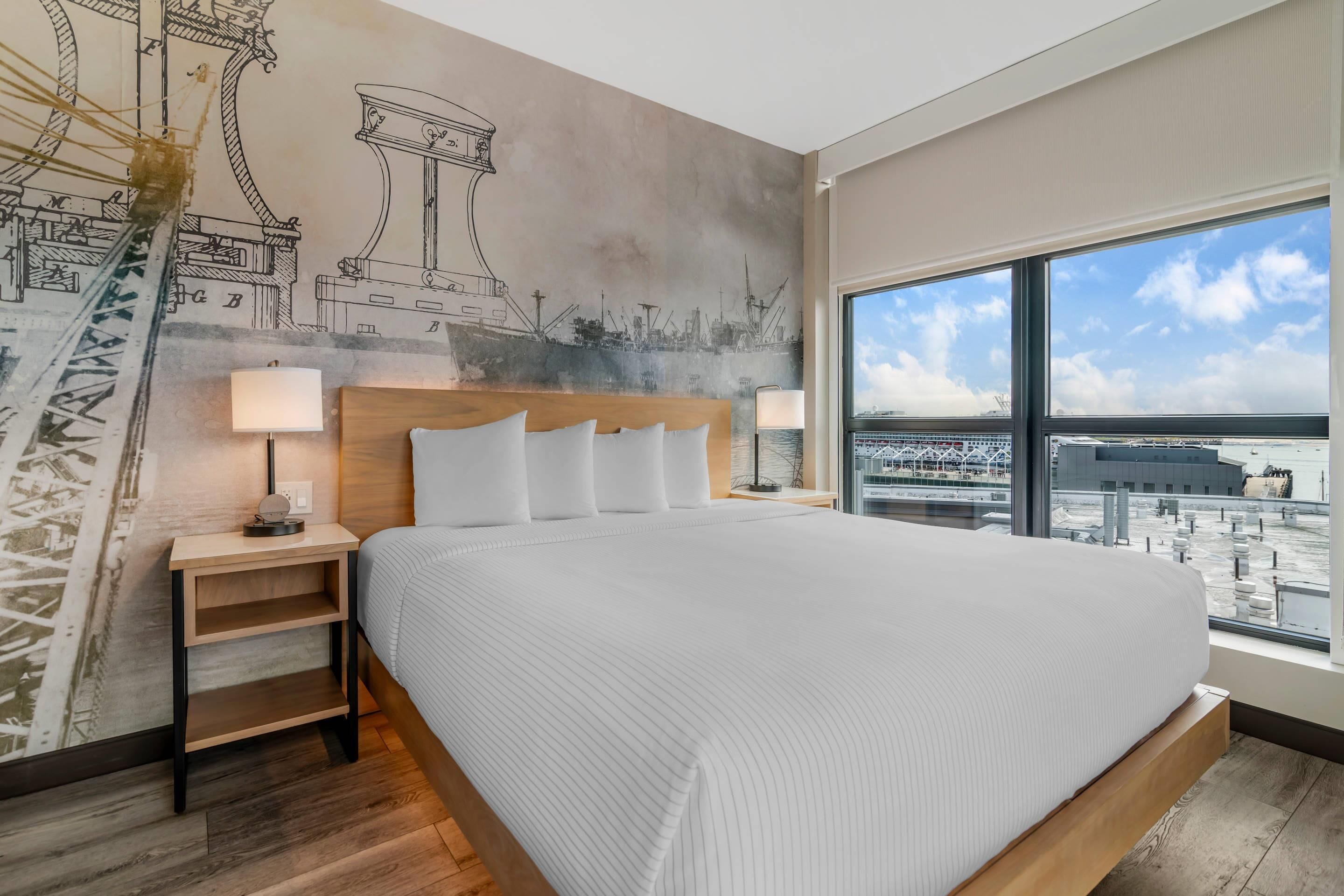 Ein modernes Hotelzimmer mit einem großen Bett, einer Stadt-Mural, einem Nachttisch und Blick auf die Stadt durch große Fenster.