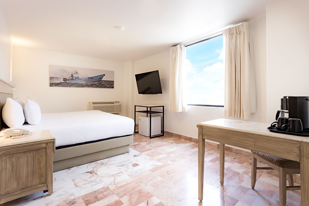Una habitación de hotel con una cama, un escritorio con cafetera, un televisor y un cuadro en la pared.