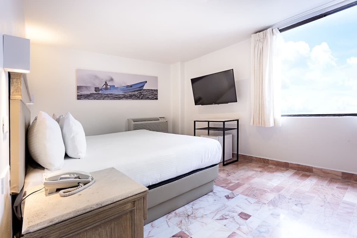 A hotel room with a bed, a bedside telephone, a flat-screen TV, a painting, and a large window.