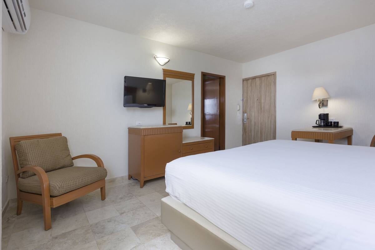 A hotel room with a bed, a wooden chair, a dresser, a television, and a lamp.