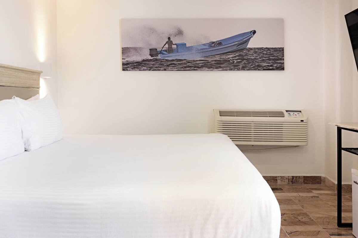 A hotel room with a bed, a wall-mounted air conditioner, and a painting of a boat on the wall.