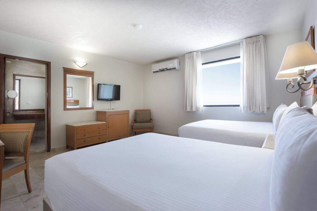 A hotel room with two beds, a television, an air conditioning unit, a desk with a chair, and a window with curtains.