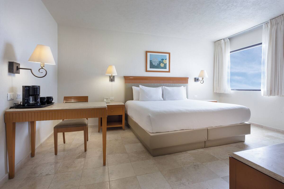 A hotel room with a bed, a desk with a chair, a coffee maker, two wall-mounted lamps, a framed picture on the wall, and a window with curtains.
