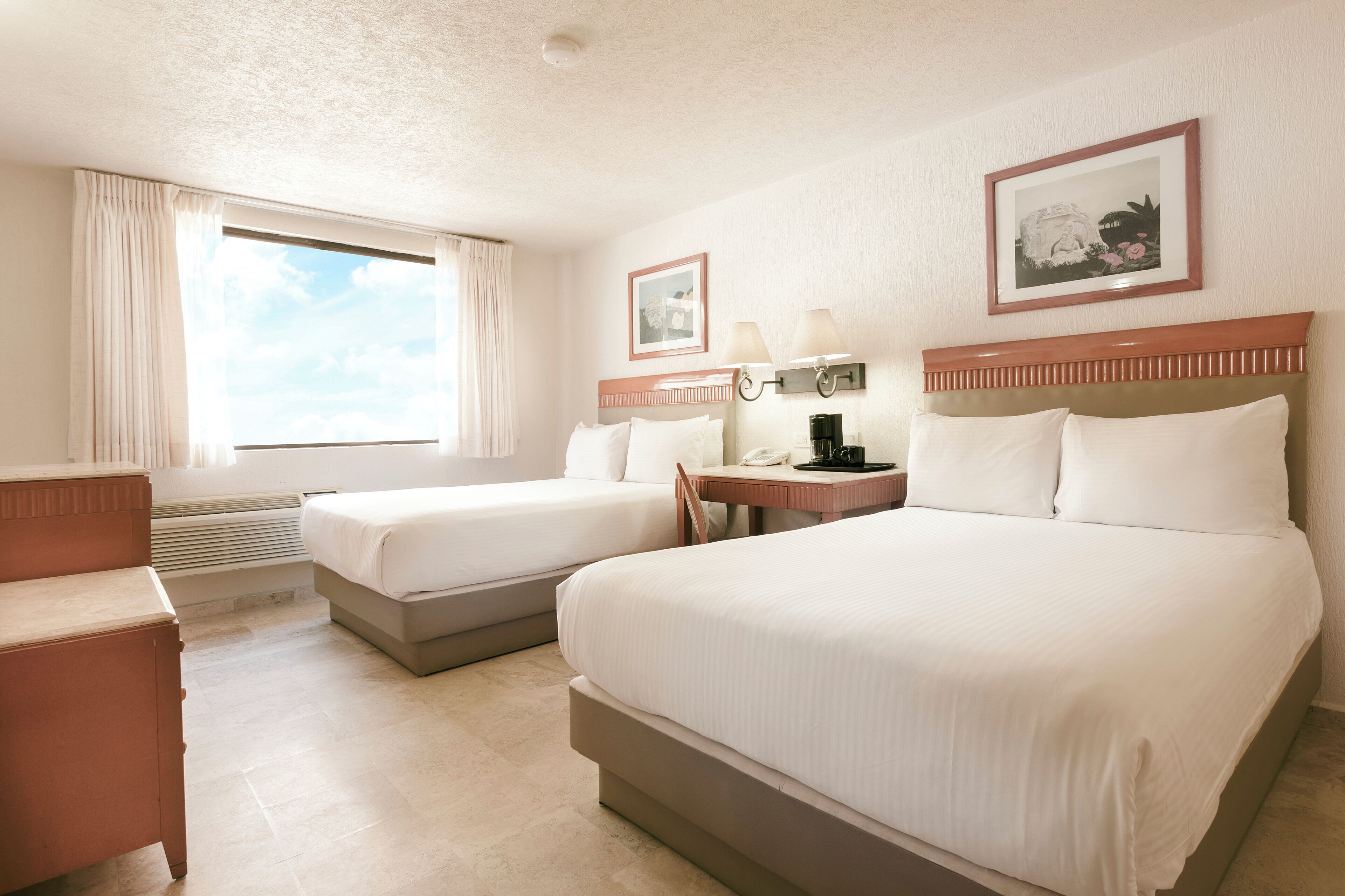 A hotel room with two beds, a desk, a chair, a coffee maker, and a window with curtains.