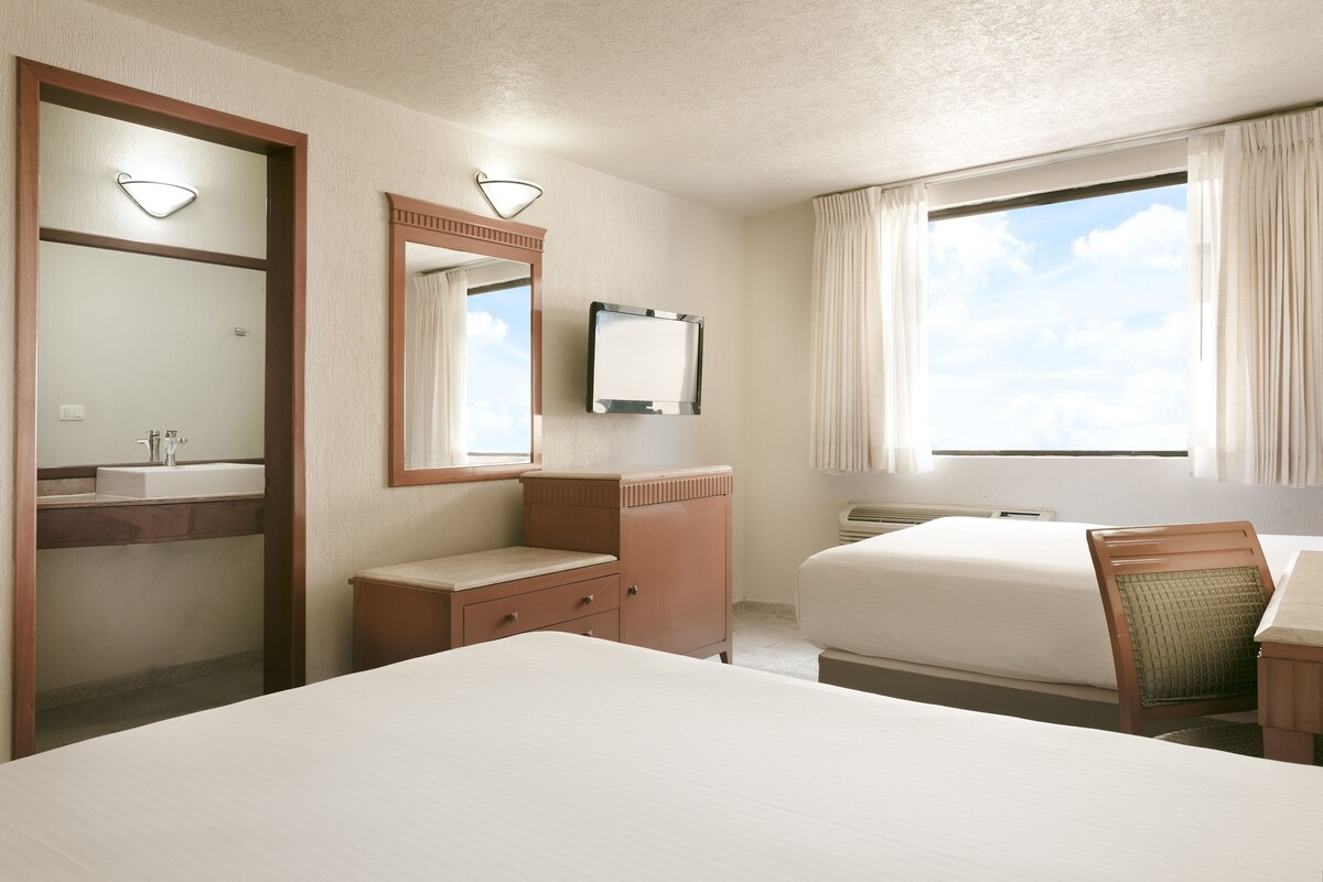 Habitación de hotel con una cama grande, una mesita de noche, una silla, un escritorio, un espejo, un televisor y una ventana con cortinas.