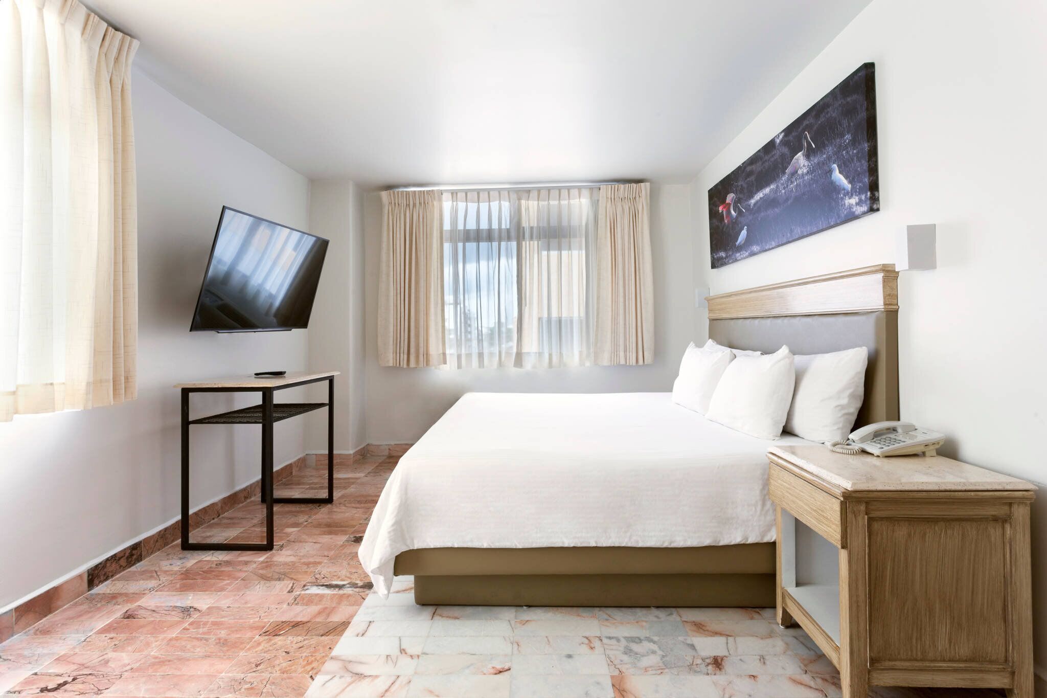 A hotel room with a bed, a TV, a bedside table, a phone, a window with curtains, and a painting on the wall.