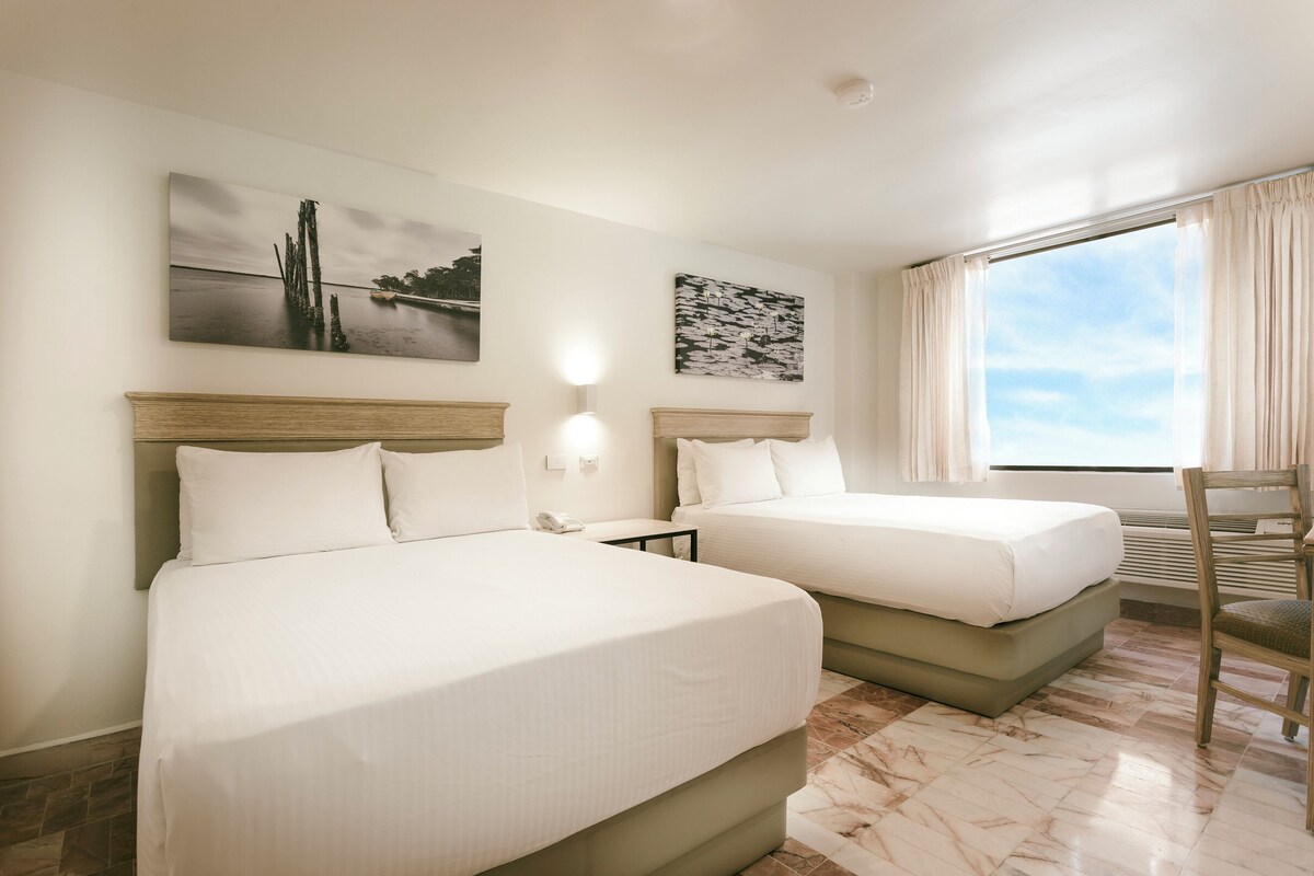 Habitación de hotel con dos camas, un ventanal con cortinas y obras de arte en las paredes.