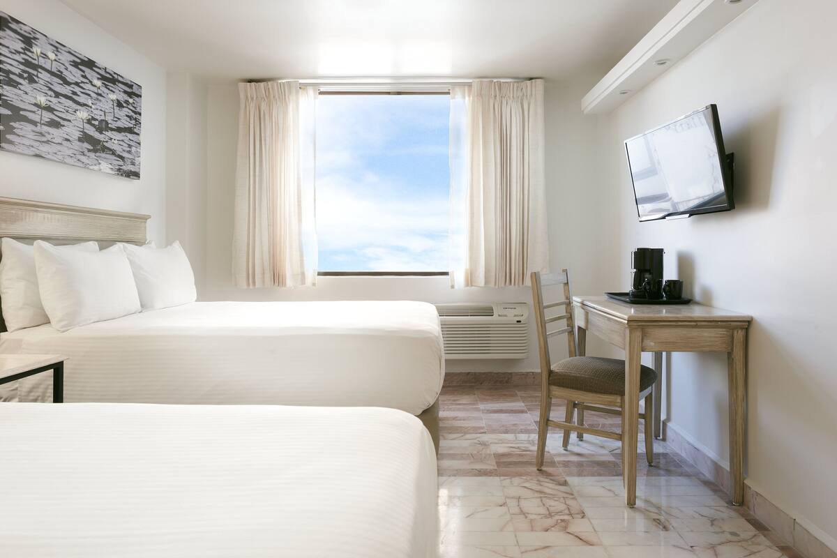 A hotel room with two beds, a desk with a chair, a flat-screen TV, and a window with curtains.