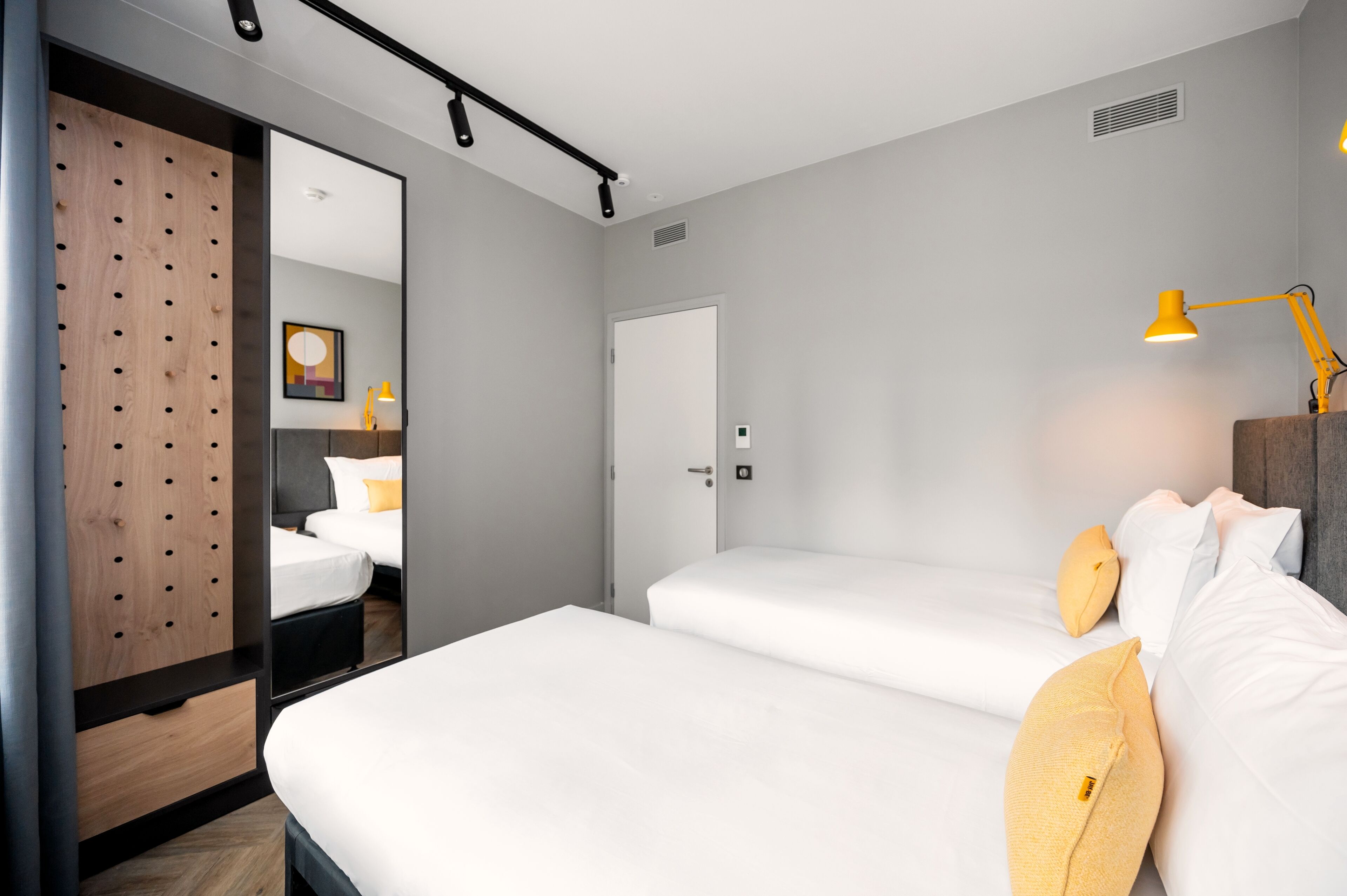 A modern hotel room with two beds, a wooden wardrobe, a mirror, and a yellow desk lamp.