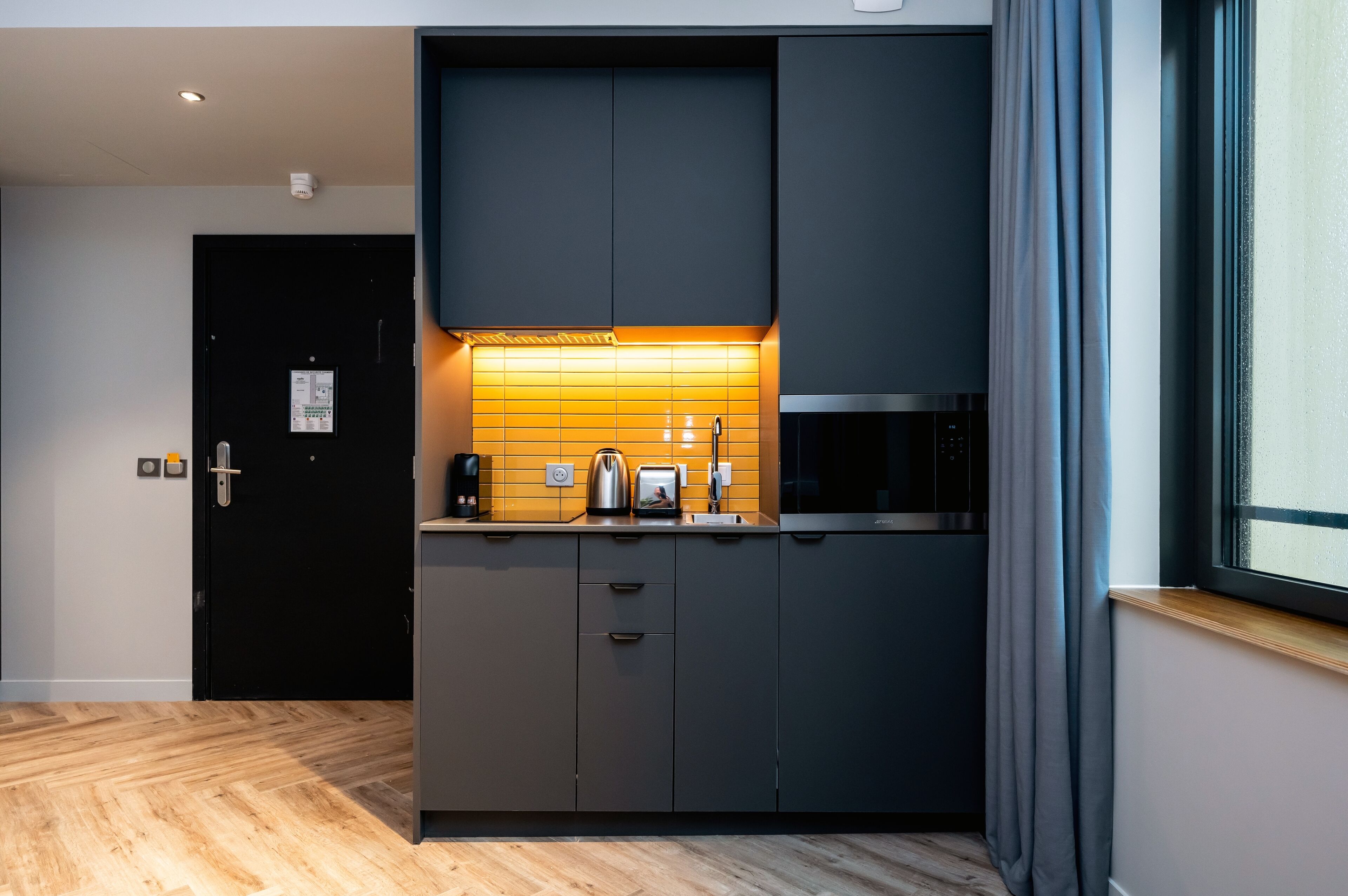 A modern kitchen with dark cabinetry, a wooden floor, and a window with curtains.