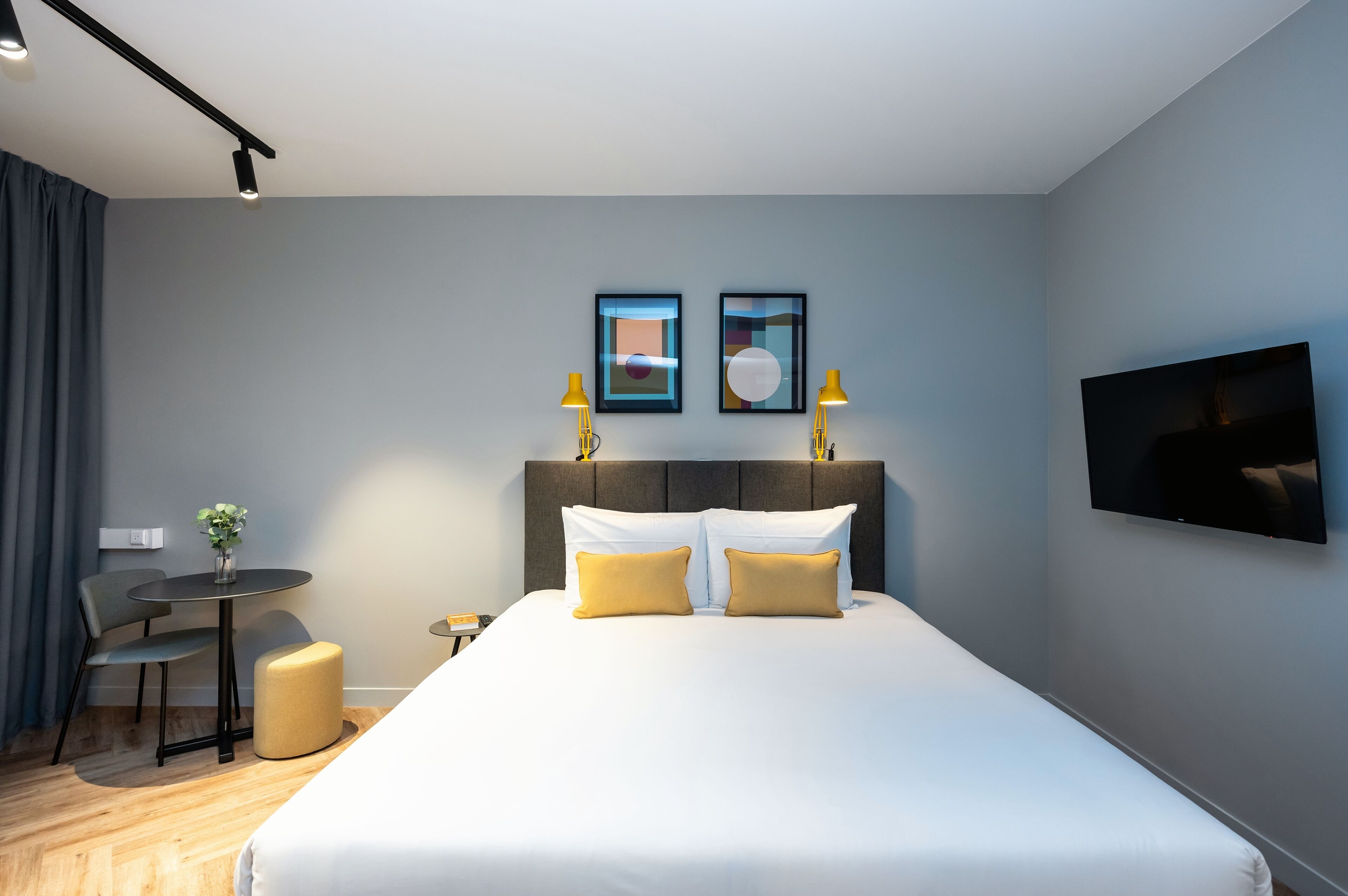 Una habitación de hotel moderna con una cama grande, un escritorio, dos sillas, una mesita con un florero y un televisor en la pared.