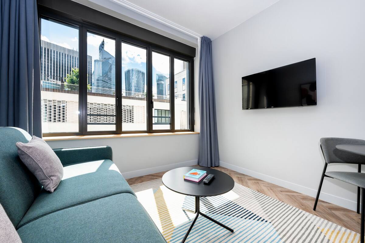 Une chambre d’hôtel moderne dotée d’une grande fenêtre, d’un canapé, d’une petite table ronde et d’un téléviseur fixé au mur.