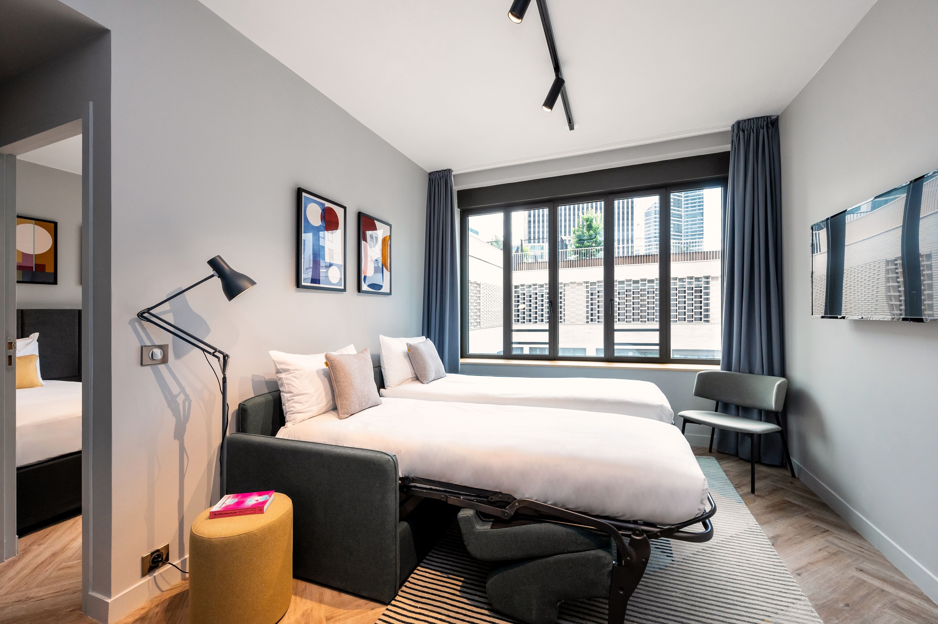 Una habitación de hotel moderna con dos camas, un escritorio, una silla, una lámpara de pie y ventanas amplias con vistas a la ciudad.