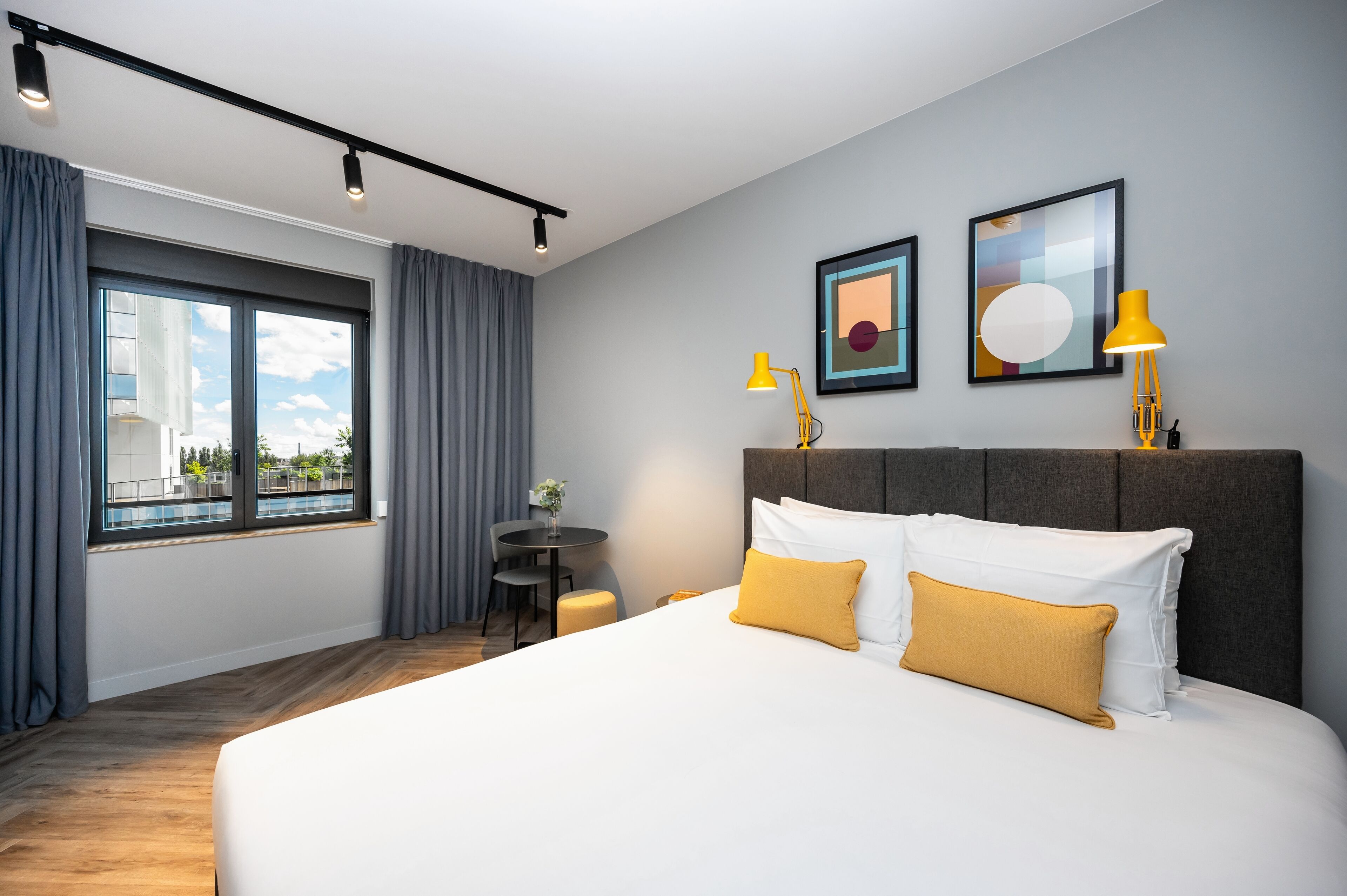 A hotel room with a large bed, two bedside lamps, a small table with a chair, and a window with curtains.