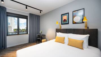A hotel room with a large bed, two bedside lamps, a small table with a chair, and a window with curtains.