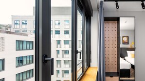 A modern hotel room with a large window, a wooden bench, a bed with a headboard, and a mirror reflecting the room.