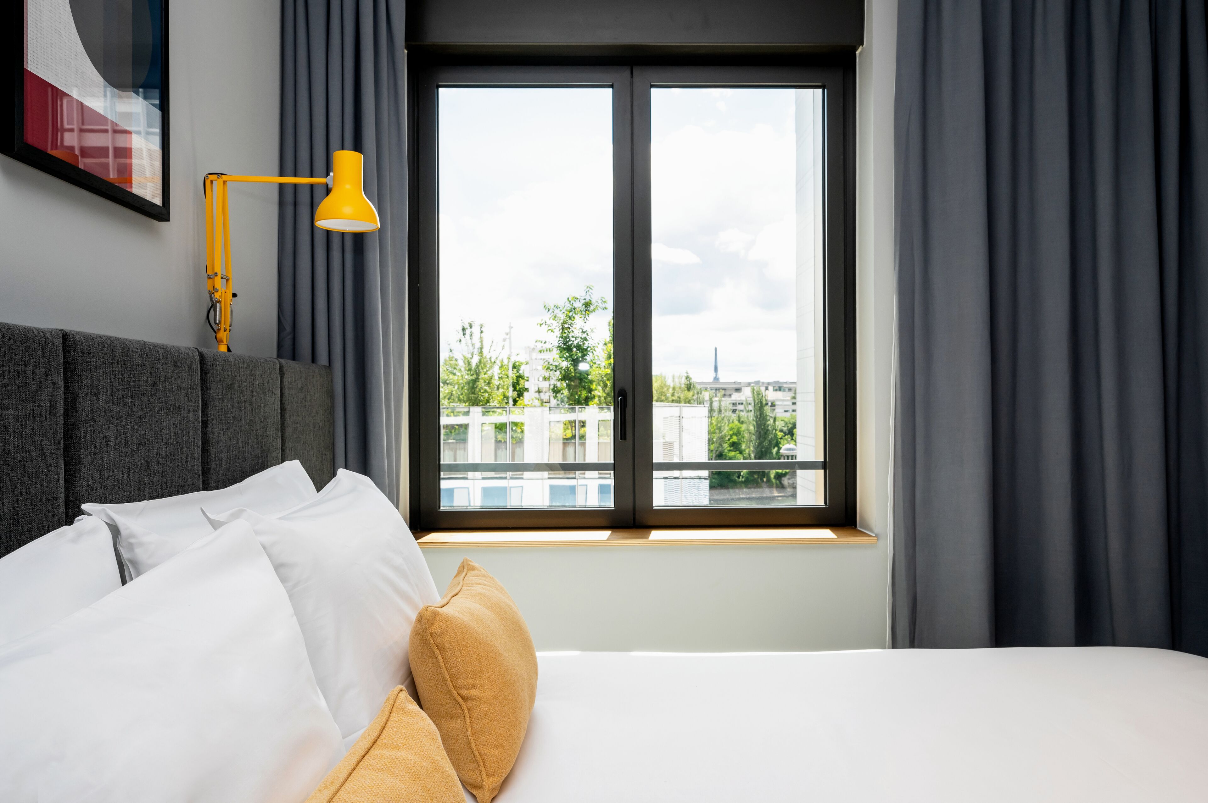 A hotel room with a bed, a window with curtains, a yellow desk lamp, and a framed picture on the wall.
