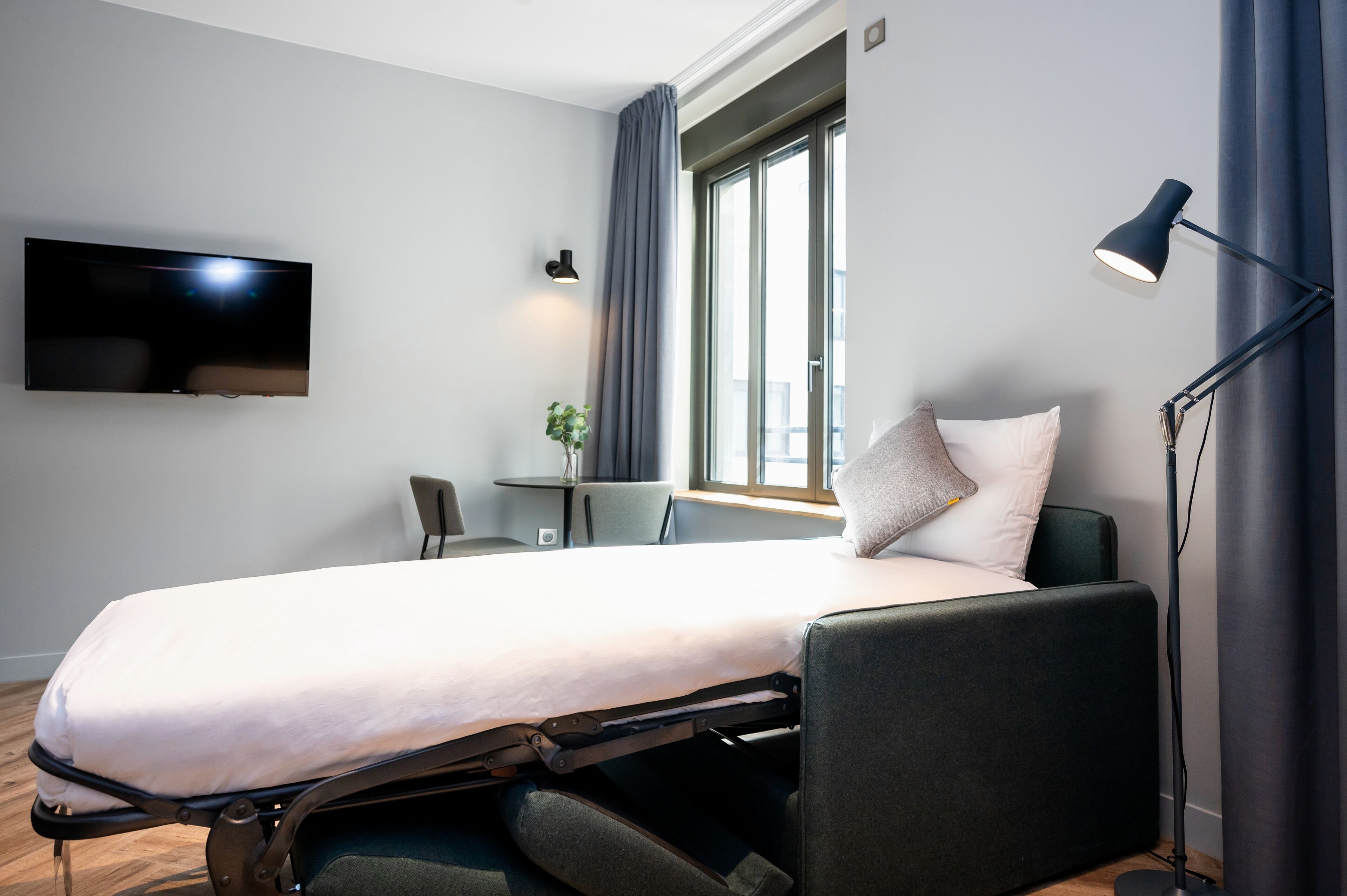 A modern hotel room with a bed, a desk, a chair, a TV, and a window with curtains.
