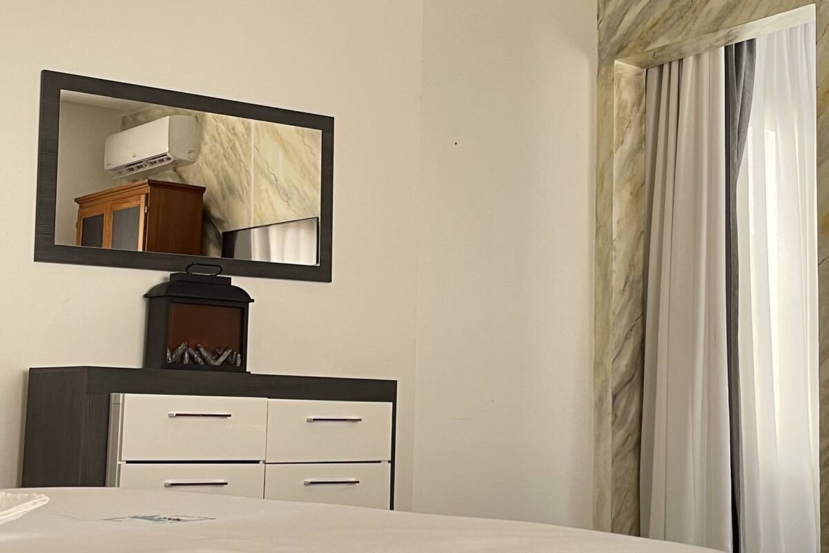 A hotel room with a bed, a dresser, a mirror, a window with curtains, and a marble wall.