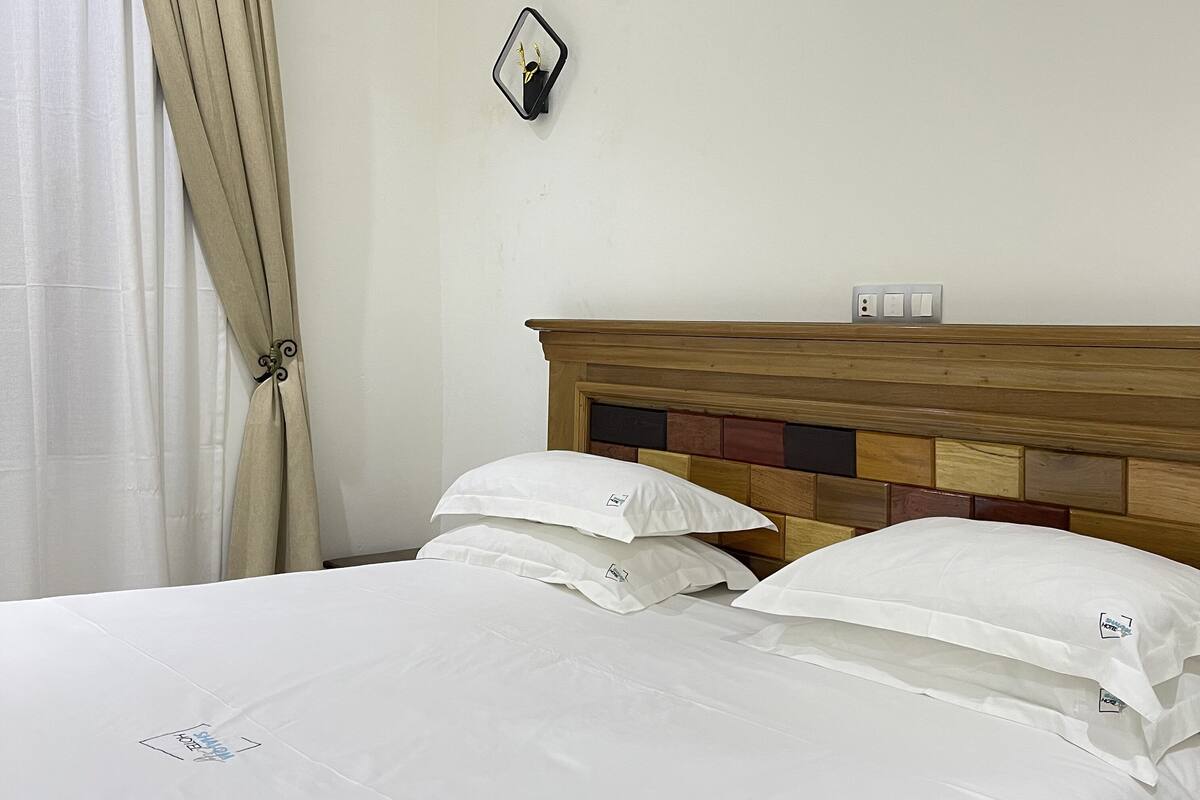 A hotel room with a wooden headboard, white bedding, an air conditioning unit, a wall-mounted mirror, and a power outlet.