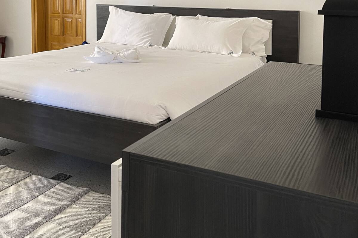 A neatly made bed with white linens, a wooden door, and a small black object on the wall.