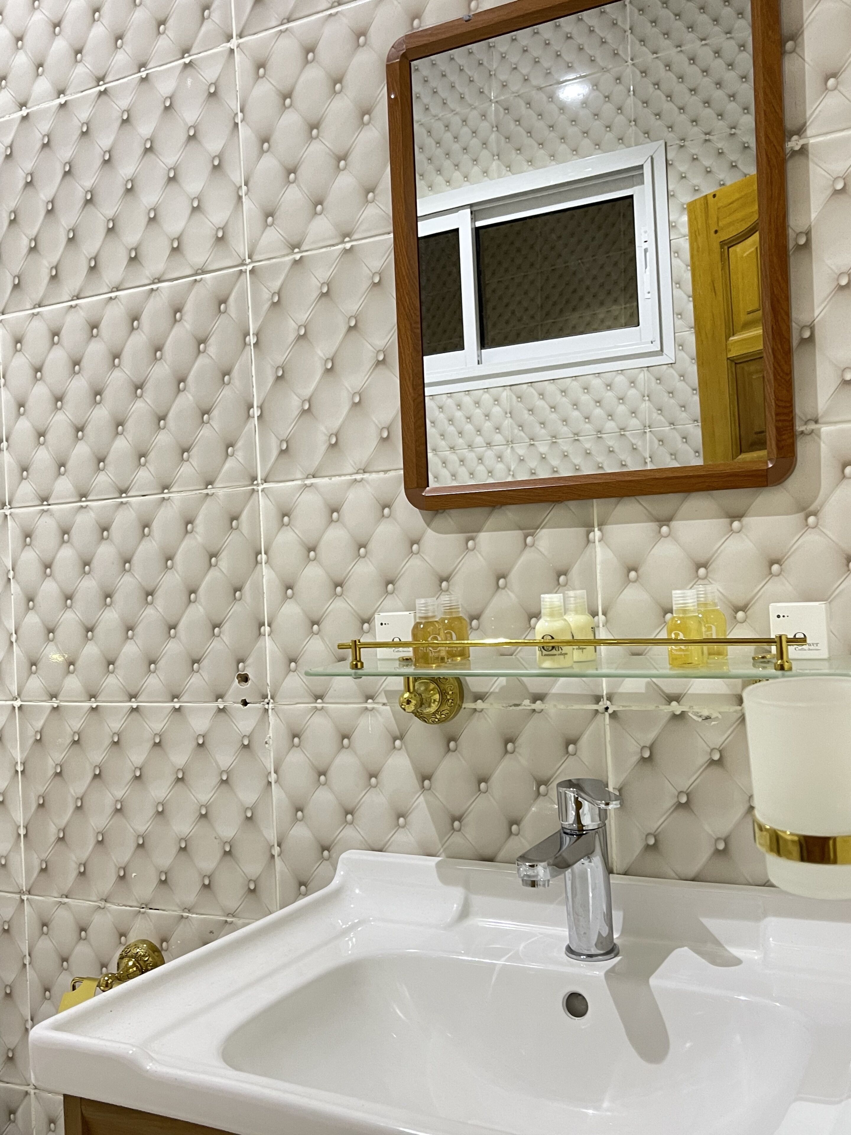 A bathroom with a tufted wall, a glass shelf with toiletries, a white sink, and a mirror with a wooden frame.