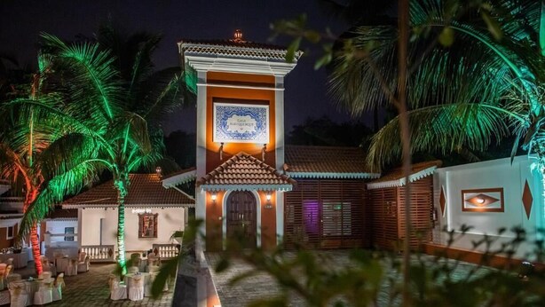 A traditional building with a tiled roof and a central tower, surrounded by palm trees and a paved courtyard. - Casa Albuquerque (Vagator)