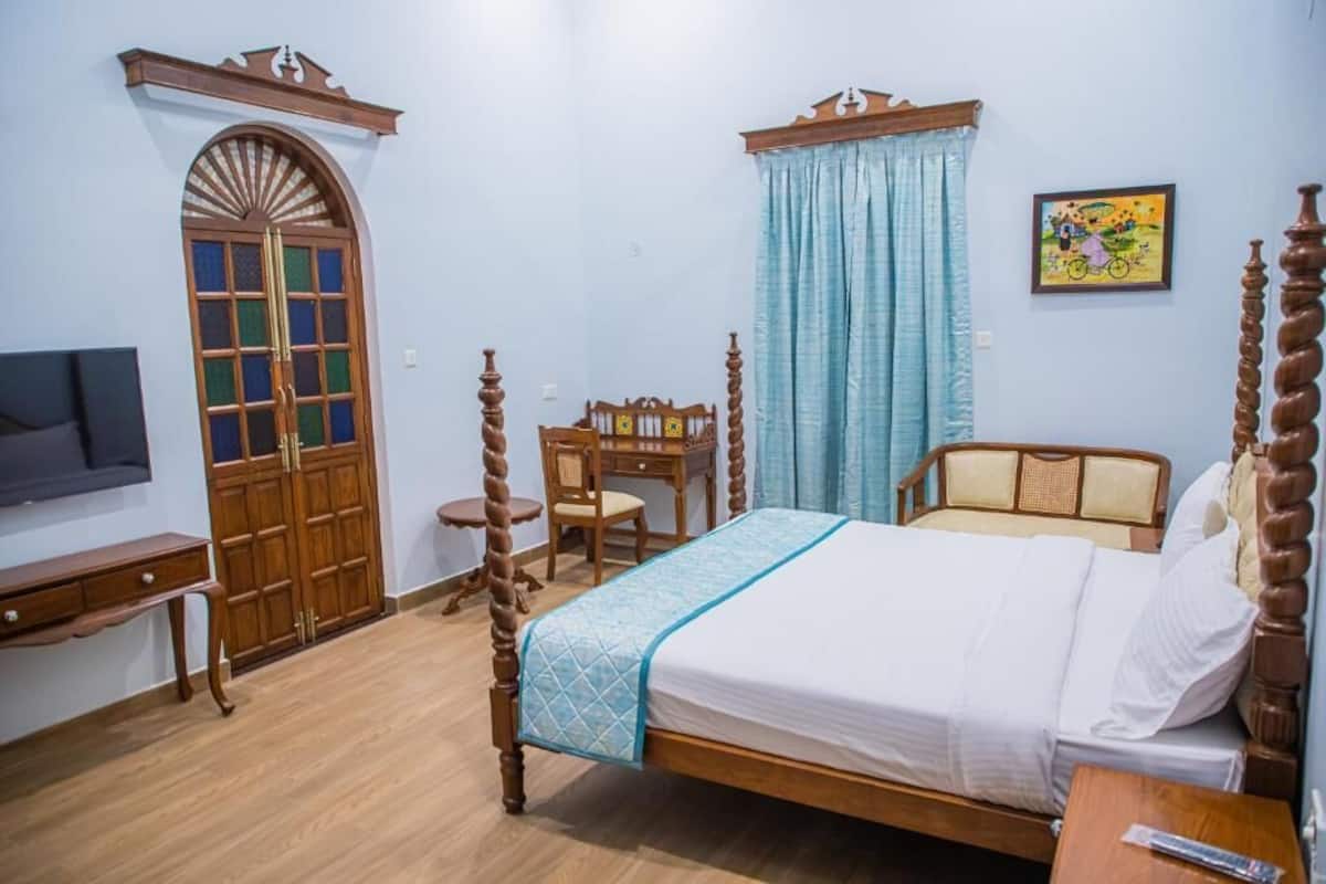 Quarto com cama de dossel de madeira, uma escrivaninha com cadeira, uma televisão e um guarda-roupa com porta de vidro.