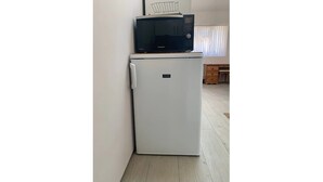 Une cuisine de taille réduite avec un réfrigérateur blanc, un micro-ondes noir posé sur le dessus, et des meubles en bois à l’arrière-plan.