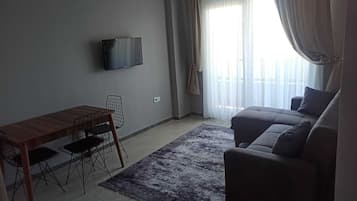 A living room with a sofa, a TV mounted on the wall, a dining table with chairs, and a large window with curtains.