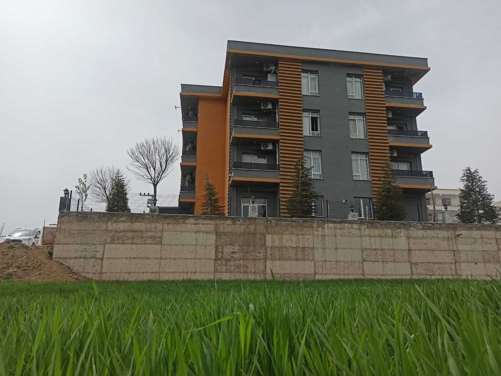 Een meerlaags woongebouw met balkons, een betonnen steunmuur en een grasveld op de voorgrond.