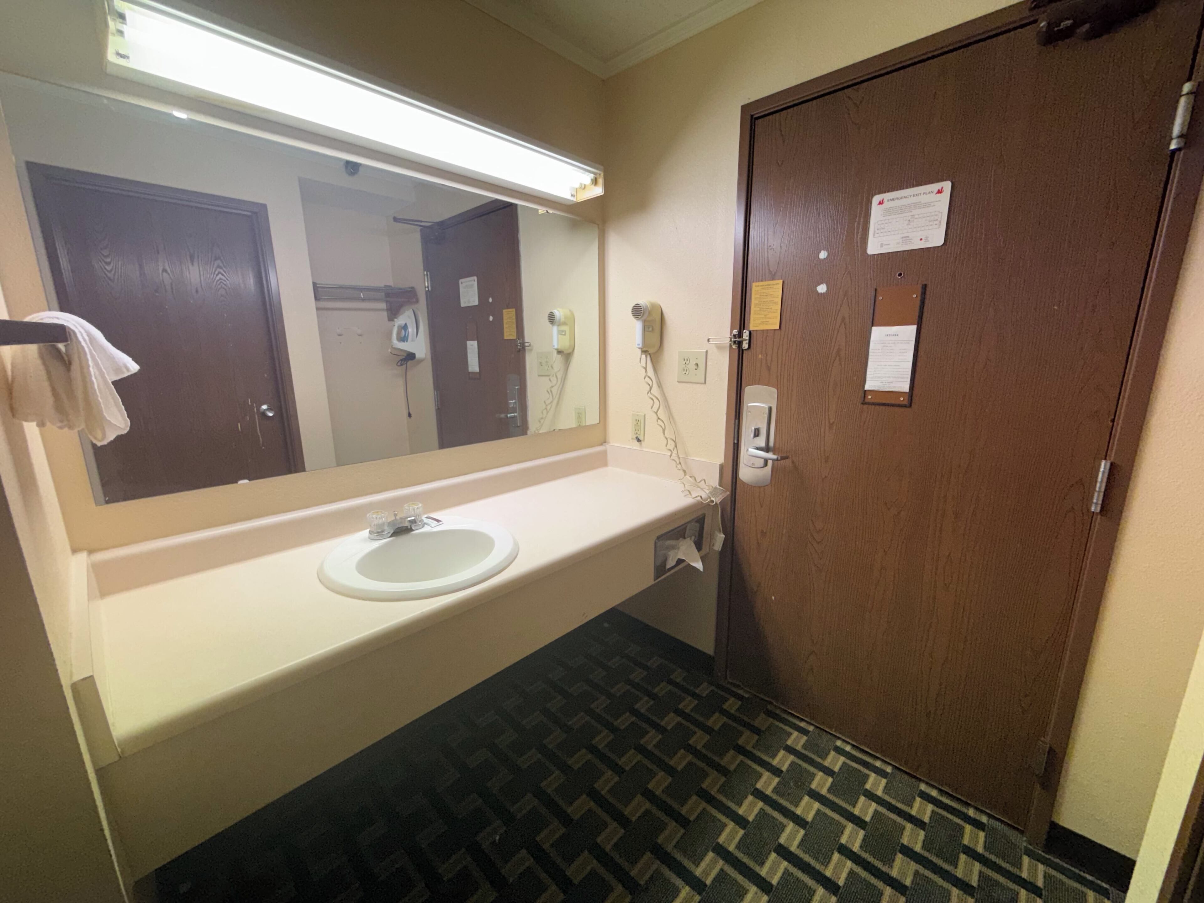 Une salle de bain avec un lavabo, un miroir et une porte dotée d’une serrure et sur laquelle est affiché un avis.