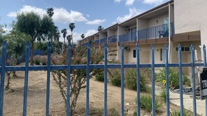 A multi-story building with balconies, surrounded by a blue fence, palm trees, and dry vegetation. - American Inn (Perris)