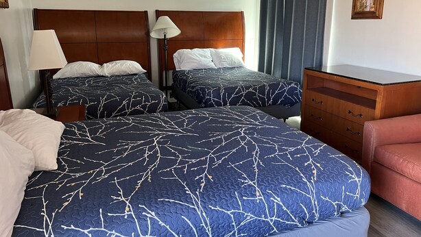 A hotel room with two beds, a wooden dresser, a desk, and a lamp. - American Inn (Perris)